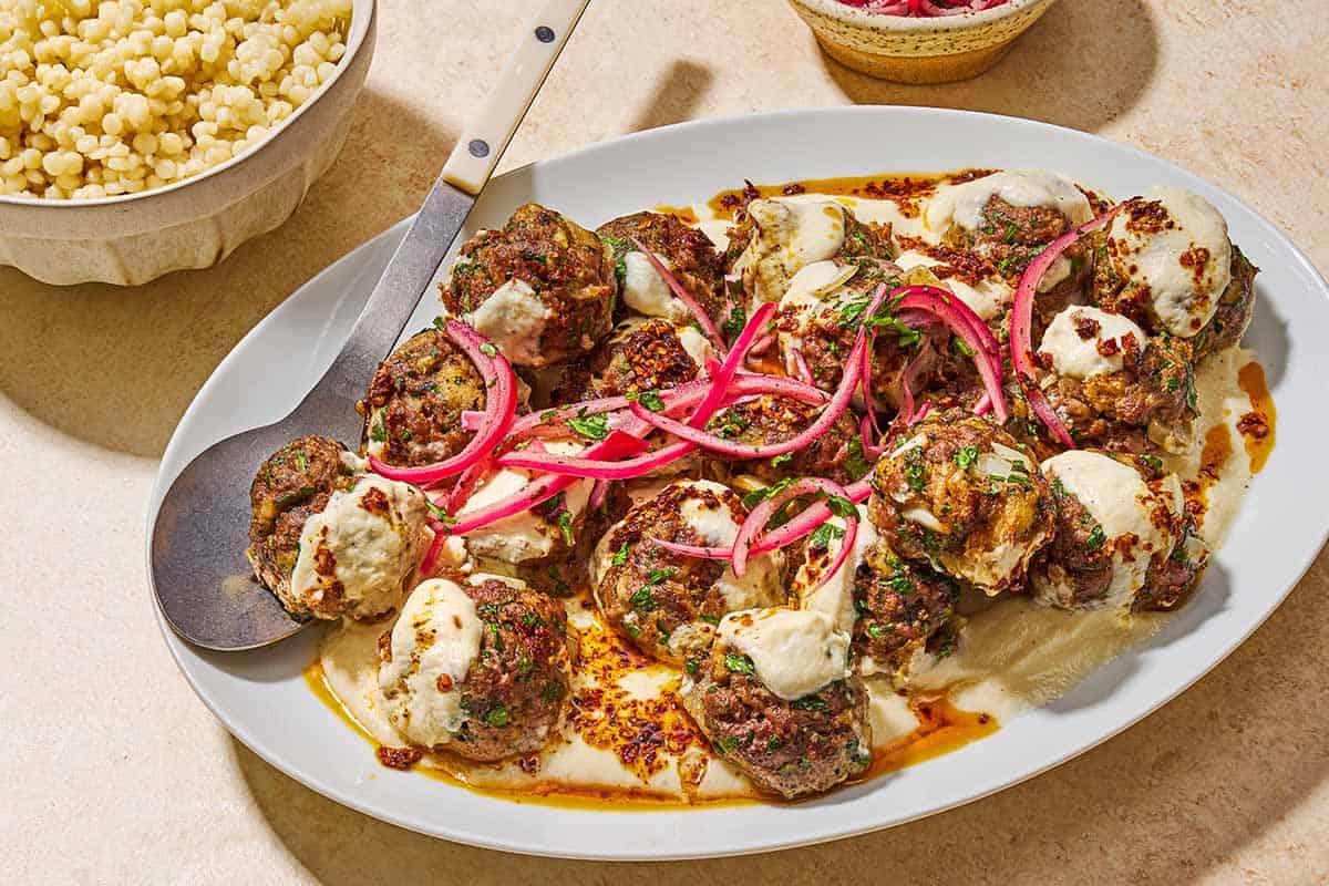 The turkish meatballs and yogurt sauce topped with pickled red onions and chopped parsley on a platter with a serving spoon next to a bowl of pearl couscous.