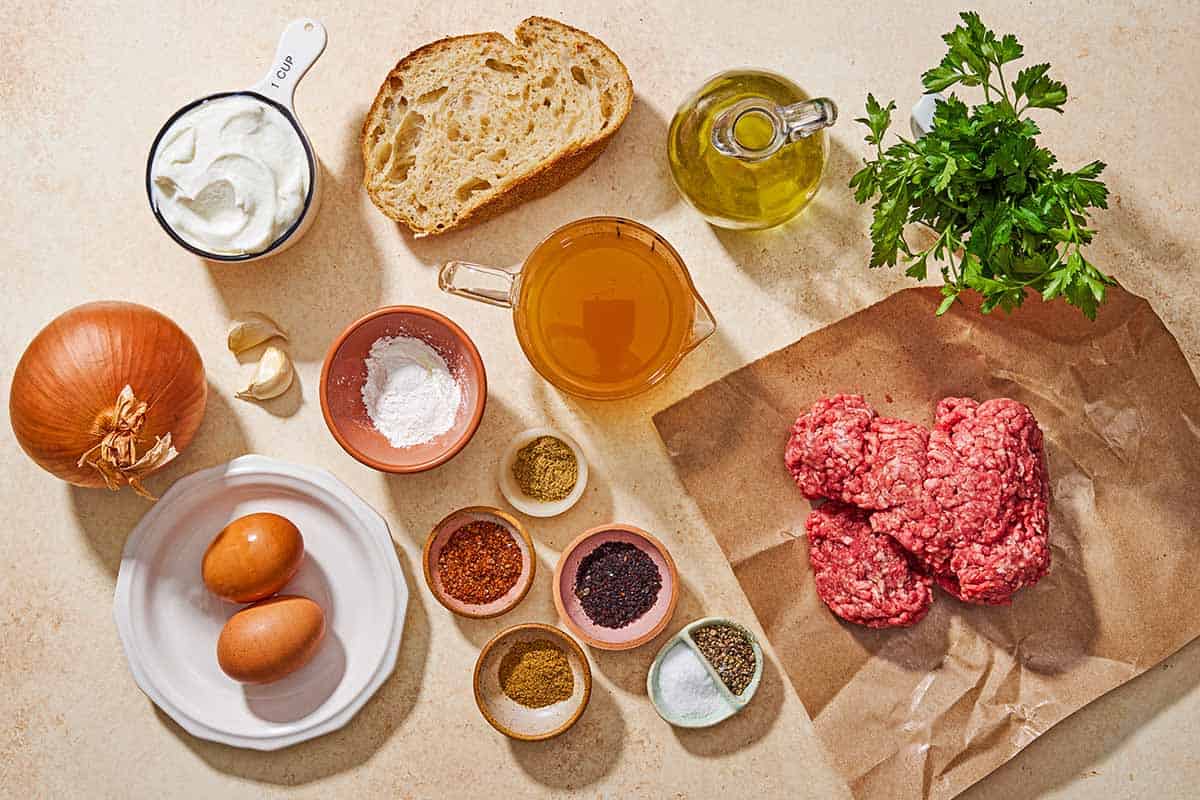 Ingredients for Turkish meatballs including a slice of bread, ground beef, onion, garlic, parsley, eggs, cumin coriander, urfa biber, aleppo pepper, salt, black pepper, cornstarch, water, greek yogurt, broth and olive oil.