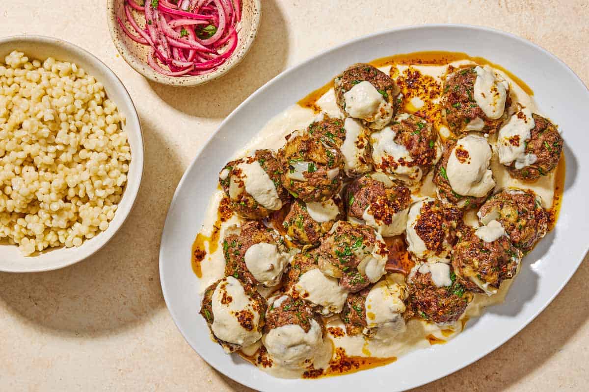 The turkish meatballs and yogurt sauce on a platter next to bowls of pearl couscous and pickled red onions.