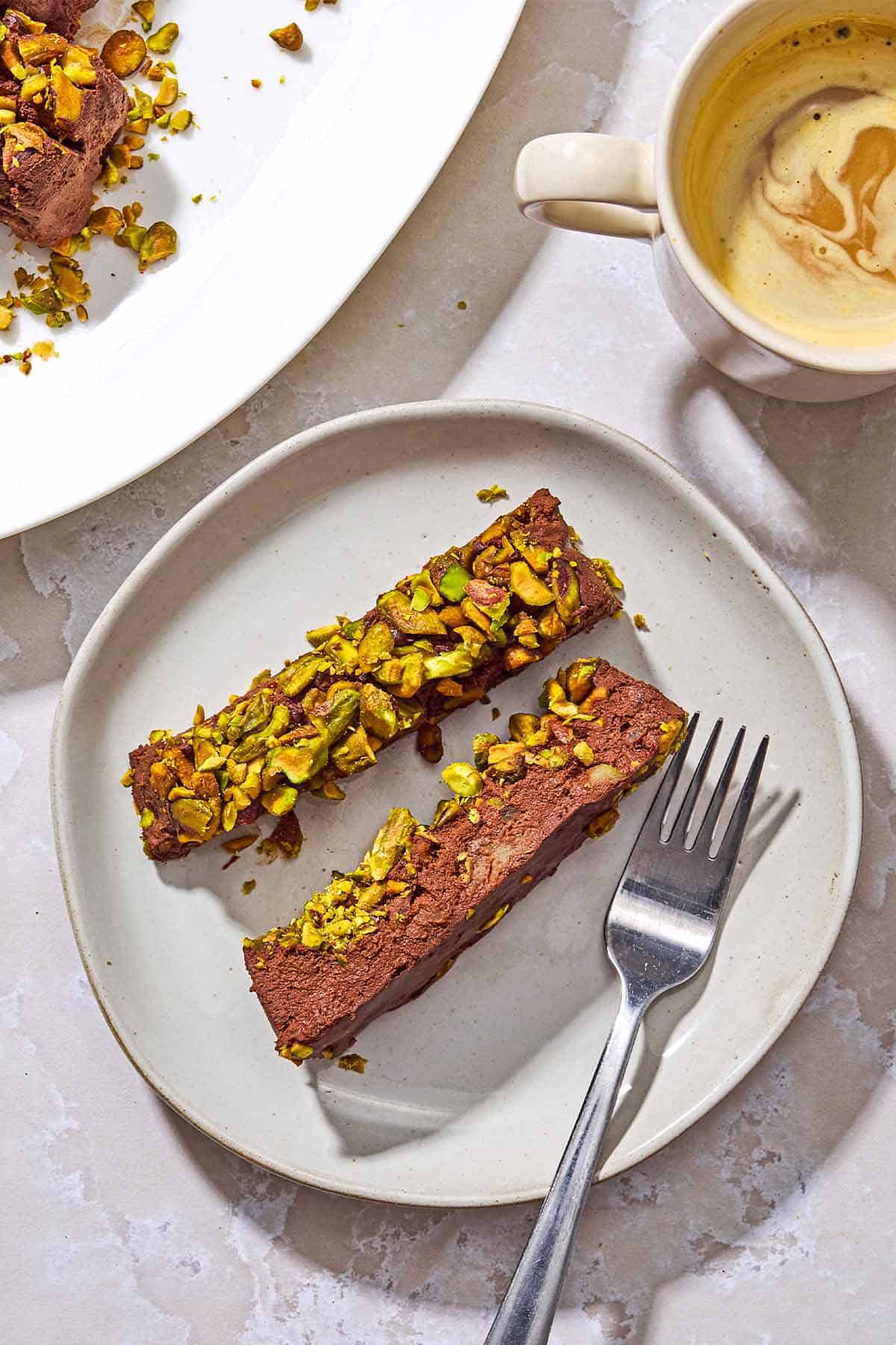 2 slices of healthy chocolate mousse on a plate with a fork next to a cup of coffee and the rest of the mousse on a platter.