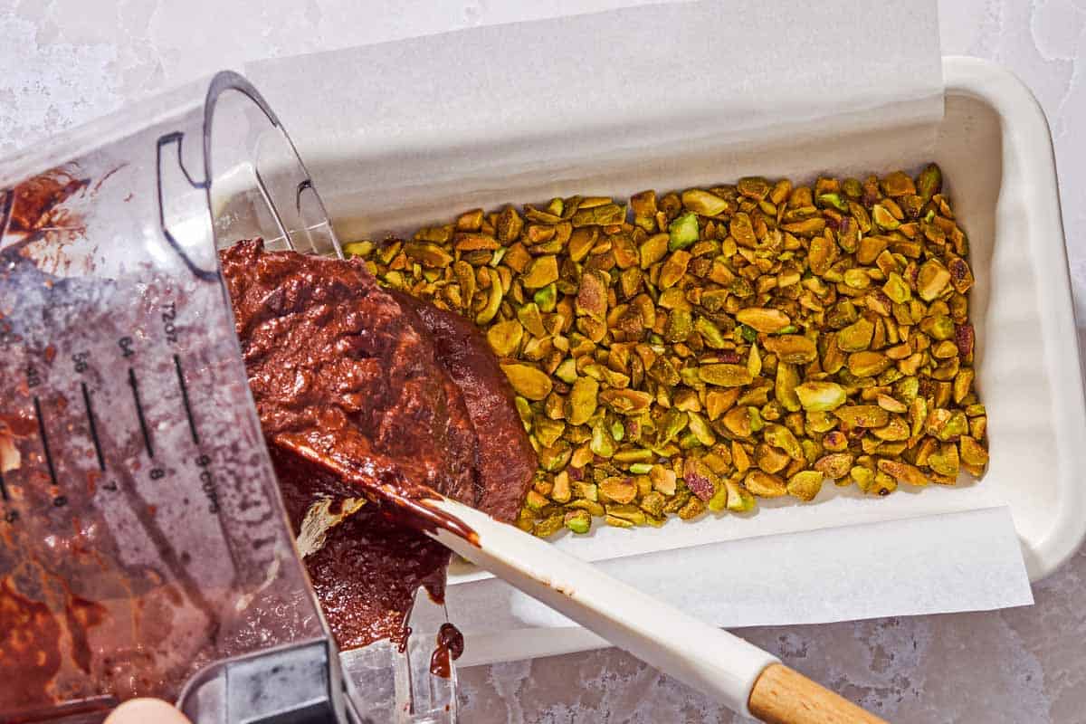 The melted chocolate and apple mixture being poured on top of pistachios in a parchment lined loaf pan.