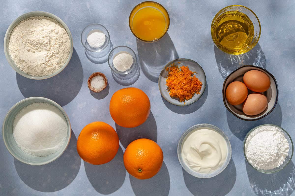 Ingredients for the orange cake recipe including oranges, eggs, sugar, greek yogurt, olive oil, orange juice, flour, baking powder, baking soda, salt, and powdered sugar.