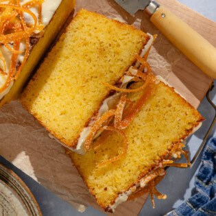 2 slices of orange cake topped with a glaze and candied orange peels next to a knife and resting in front of the rest of the loaf. Surrounding this is a plate of orange wedges, a kitchen towel and a stack of 2 plates.