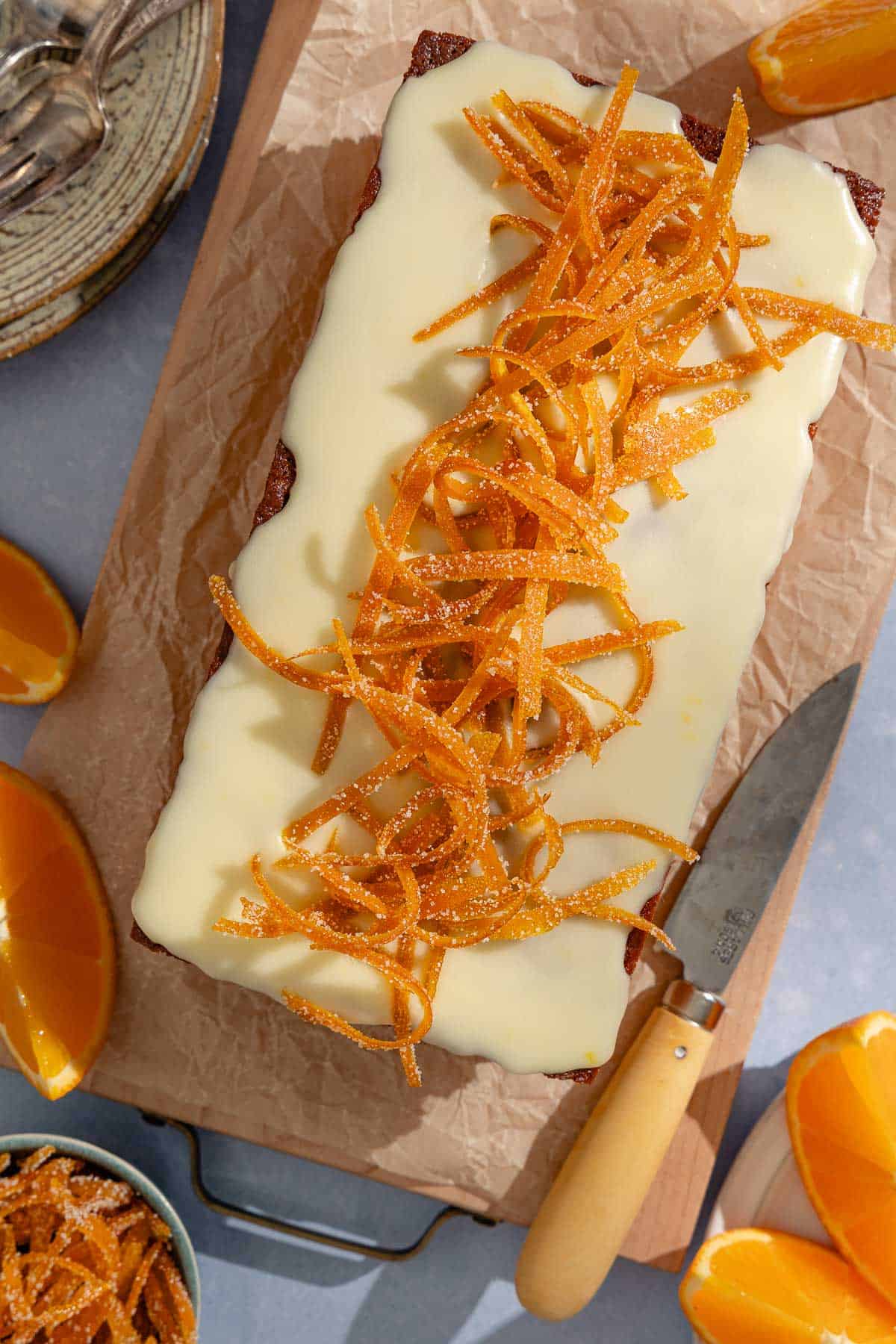 An orange cake topped with glaze and candied orange peels sitting on a parchment lined cutting board with a knife and orange wedges. Next to this is a bowl of candied orange peels, more orange wedges, and a stack of plates with forks.