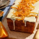 Orange cake topped with a glaze and candied orange peels sitting on a parchment lined cutting board surrounded by orange wedges and vrious other table settings.