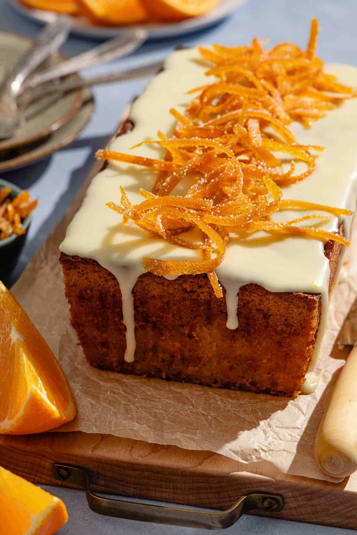 Orange cake topped with a glaze and candied orange peels sitting on a parchment lined cutting board surrounded by orange wedges and vrious other table settings.