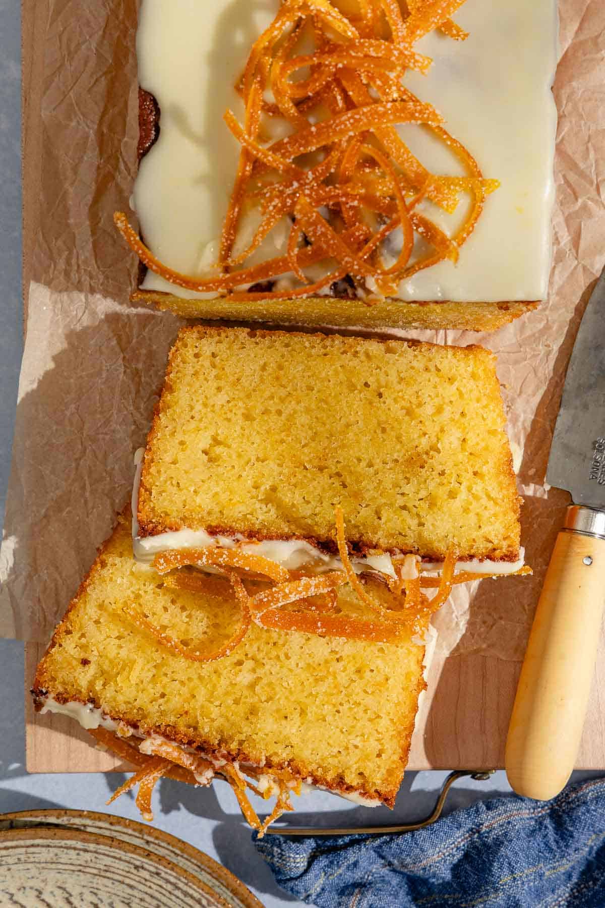 2 slices of orange cake topped with a glaze and candied orange peels next to a knife and resting in front of the rest of the loaf.