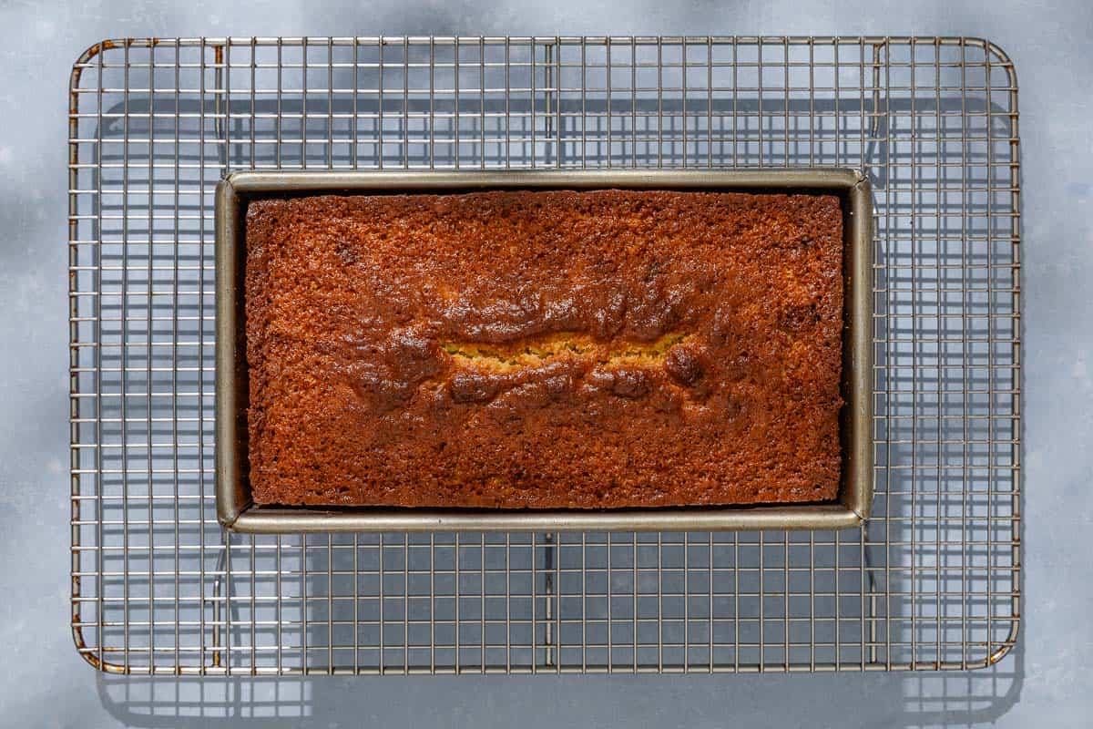 A baked orange cake in a loaf pan cooling on a wire rack.