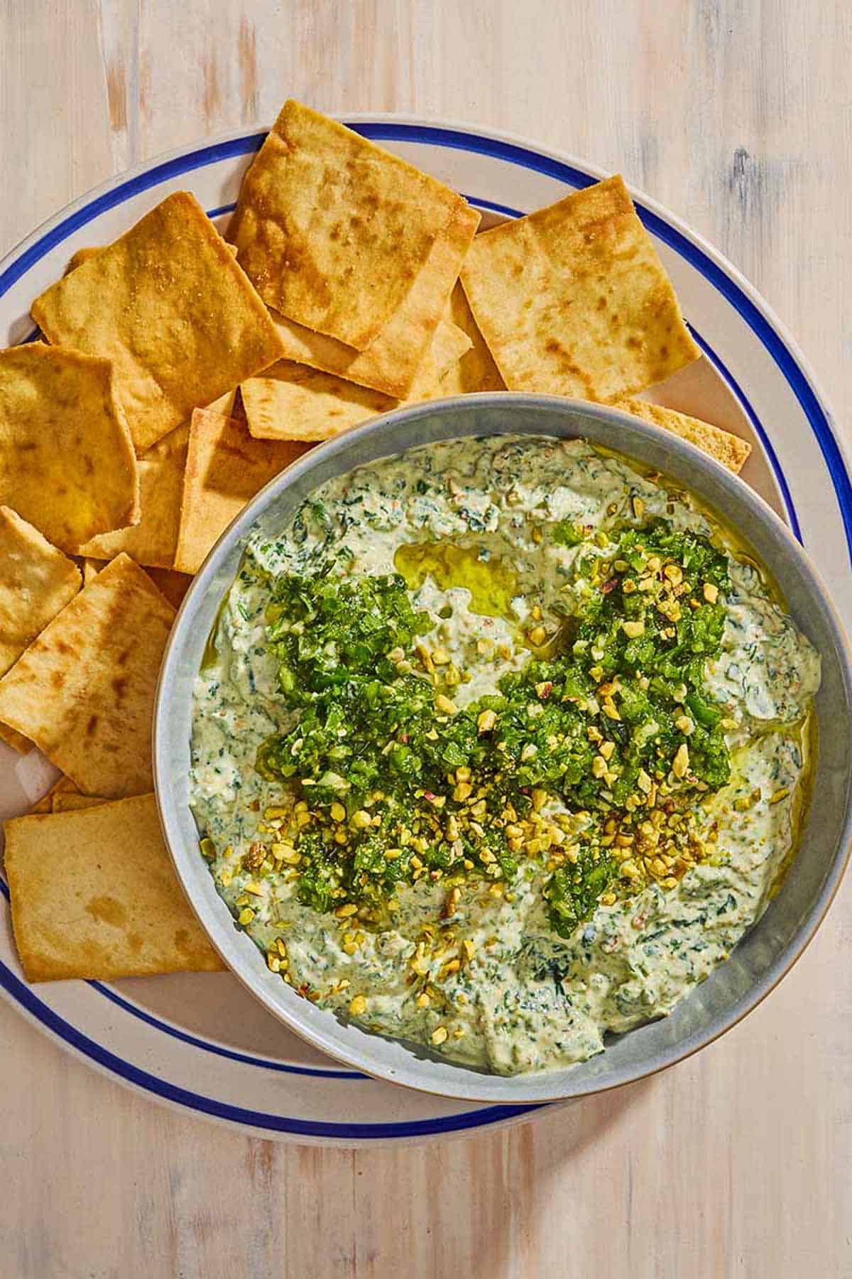 A bowl of spinach dip topped with pistachios and zhoug on a plate with pita chips.