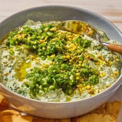 Spinach dip topped with pistachios and zhoug in a bowl with a spoon on a plate with pita chips.