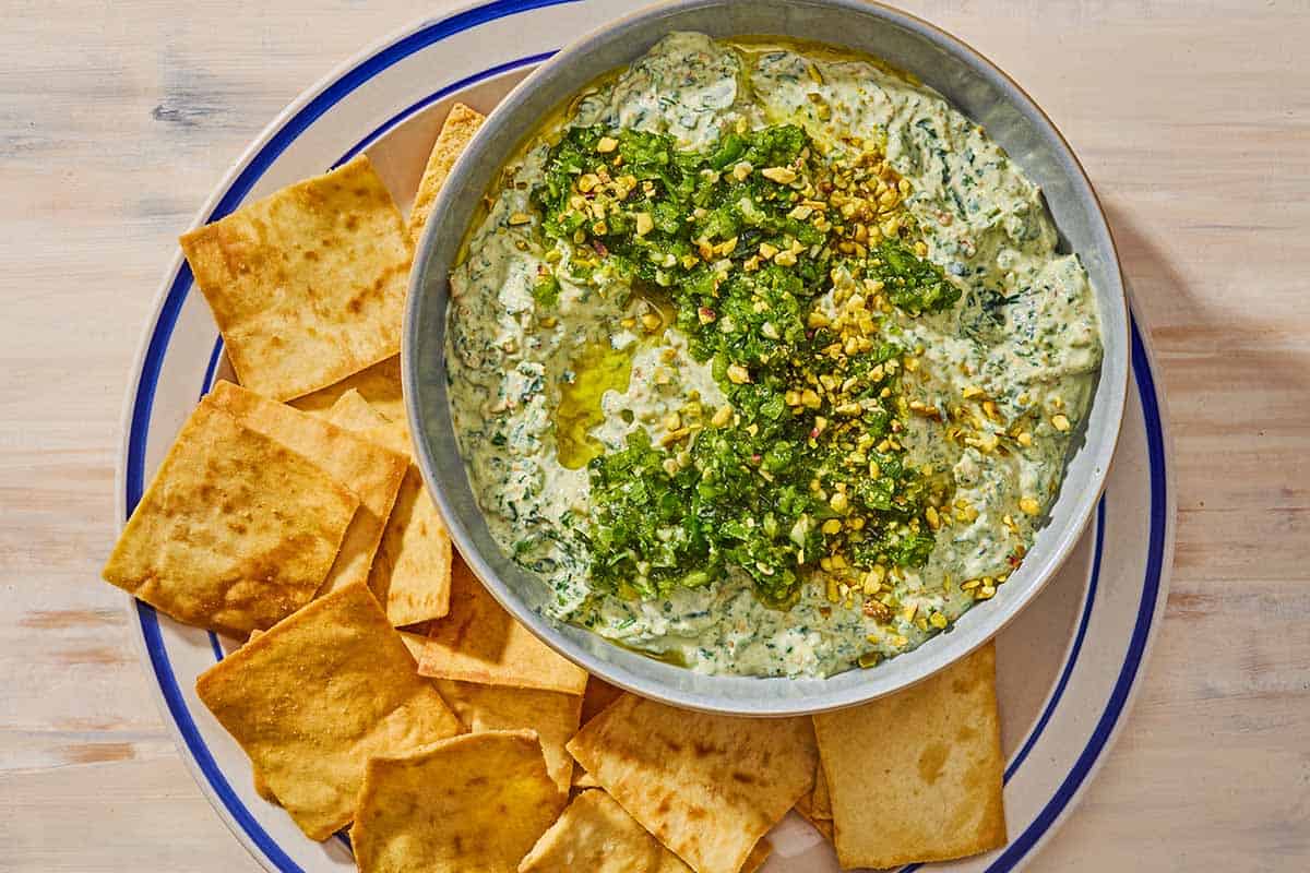 A bowl of spinach dip topped with pistachios and zhoug on a plate with pita chips.