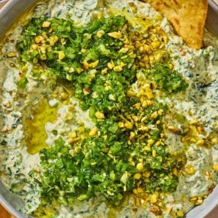 A bowl of spinach dip topped with pistachios and zhoug on a plate with pita chips.